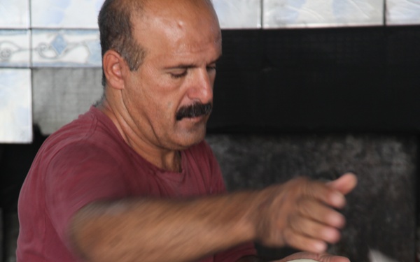 A middle-aged man wearing a red t-shirt is working in a bakery, actively stretching or placing flatbread dough onto what appears to be a conveyor or baking tray leading toward an oven. His arms are in motion, indicating active bread-making work. The setting appears to be a traditional Middle Eastern or Mediterranean bread bakery.
