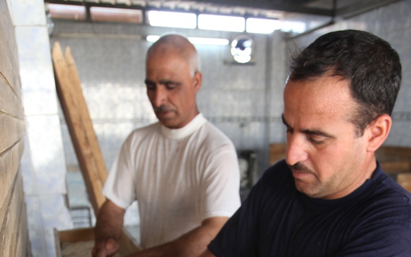 Two men are working in a bakery, handling flat bread dough in a long wooden trough or proofing box. The man in the foreground wears a dark navy t-shirt and is arranging raw flatbread dough pieces, while the man in the background wears a white t-shirt and appears to be using a wooden paddle or peel. The setting appears to be a traditional or artisanal flatbread bakery with tiled walls.