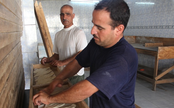 Two men are working in what appears to be a traditional flatbread bakery. The man in the foreground wearing a navy blue shirt is handling flatbread dough in a wooden trough, while the man in the background wearing a white t-shirt assists with a wooden board. The setting is a tiled room with wooden work surfaces consistent with a Middle Eastern or Mediterranean bread-making operation.