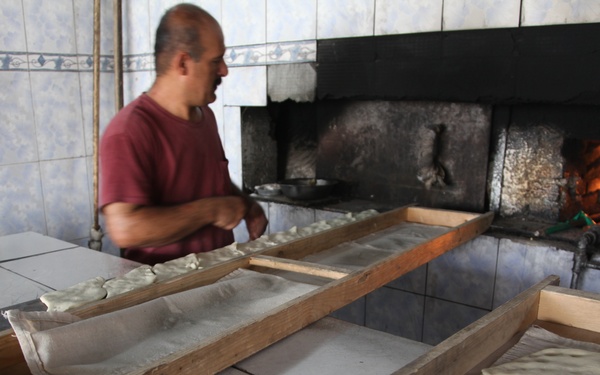 A man works in a traditional bakery, preparing flatbread dough on long wooden-framed trays. The dough pieces are shaped and laid out on the trays, ready to be baked in a wood-fired oven visible in the background. The setting appears to be a small artisan bread-making operation with tiled walls and a heavily soot-stained oven opening.