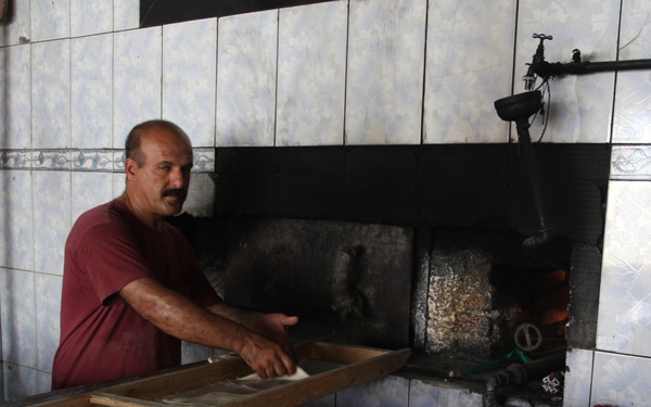 A man in a red shirt is working in a traditional bakery, loading or retrieving flatbread dough into a large wood or gas-fired oven. The workspace features blue and white tiled walls, wooden bread-proofing or cooling racks in the foreground holding shaped dough, and industrial piping along the walls. The setting appears to be a Middle Eastern or Central Asian bread bakery.
