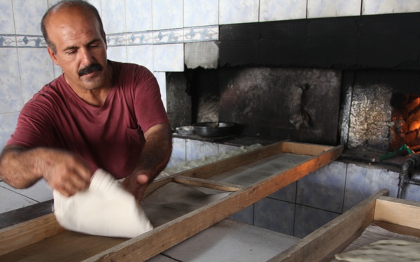 A middle-aged man with a mustache is working in a traditional bakery, shaping or tossing raw dough on a wooden-framed tray. Several shaped dough pieces are already lined up in a parallel tray, ready for baking. A wood-fired oven with visible flames burns in the background.