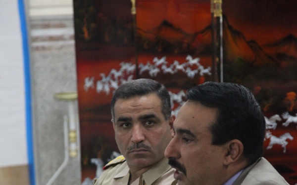 Two men are seated at a wooden table in what appears to be an official meeting or briefing setting. One man is wearing a desert camouflage military uniform with insignia patches, while the other is dressed in a civilian suit with a striped tie. An open binder or document folder is visible on the table in front of them.