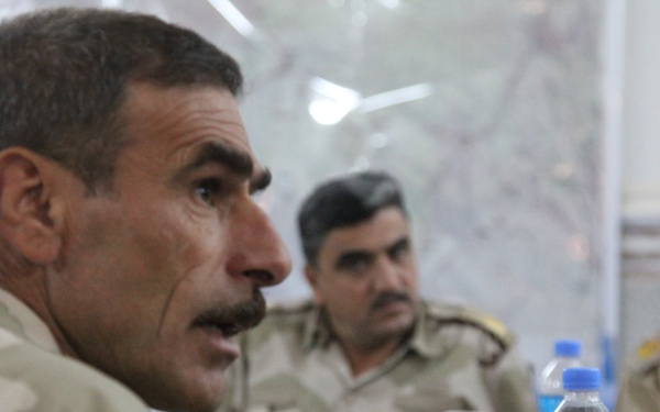 Several military personnel in desert camouflage uniforms are seated around a table, apparently engaged in a meeting or briefing. The foreground figure appears to be speaking, while others listen in the background. Beverages including water bottles and canned drinks are visible on the table.