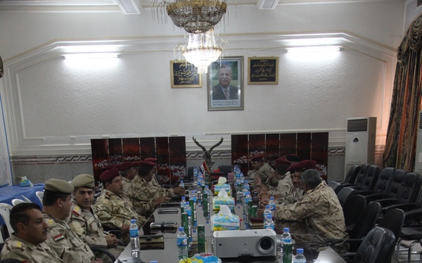 A formal military briefing or meeting is taking place in an ornate conference room. Multiple uniformed military personnel are seated around a large conference table, with some wearing red berets and others in camouflage uniforms. The room features framed portraits on the wall, a chandelier, and a projector on the table.