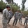 A group of five U.S. Army soldiers in ACU (Army Combat Uniform) digital camouflage are gathered outdoors in what appears to be a Middle Eastern environment. One soldier, facing away from the camera, appears to be briefing or instructing the others while they look down at a disturbed patch of ground. The setting includes sandy/gravelly terrain, palm trees, and a concrete wall in the background.