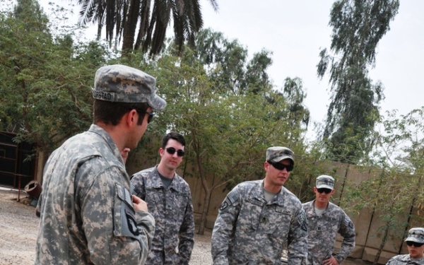 A group of five U.S. Army soldiers in ACU (Army Combat Uniform) digital camouflage are gathered outdoors in what appears to be a Middle Eastern environment. One soldier, facing away from the camera, appears to be briefing or instructing the others while they look down at a disturbed patch of ground. The setting includes sandy/gravelly terrain, palm trees, and a concrete wall in the background.