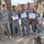 A group of military personnel pose together outdoors, with several individuals holding certificates of training. The group includes soldiers in US Army ACU (Army Combat Uniform) pattern and others in blue digital camouflage uniforms, likely Iraqi Security Forces. The setting appears to be a forward operating base or military compound with camouflage netting visible in the background.