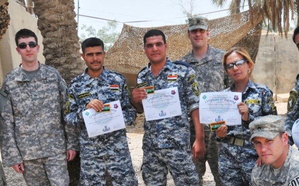 A group of military personnel pose together outdoors, with several individuals holding certificates of training. The group includes soldiers in US Army ACU (Army Combat Uniform) pattern and others in blue digital camouflage uniforms, likely Iraqi Security Forces. The setting appears to be a forward operating base or military compound with camouflage netting visible in the background.