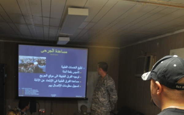 A military briefing or training session is taking place in what appears to be a forward operating base or military facility. A presentation with Arabic text is being displayed on a projection screen and mirrored on a laptop, suggesting a joint or coalition training environment. Personnel in both military uniforms and civilian/Army-branded clothing are present as attendees and presenters.