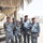 A group of six uniformed personnel wearing blue digital camouflage uniforms pose together outdoors on a gravel surface. They are standing around a tracked ground robot, consistent with an EOD (Explosive Ordnance Disposal) remotely operated vehicle. Two individuals wear red berets, and at least one uniform displays an 'EOD' patch, indicating a bomb disposal unit context.