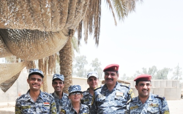 A group of six uniformed personnel wearing blue digital camouflage uniforms pose together outdoors on a gravel surface. They are standing around a tracked ground robot, consistent with an EOD (Explosive Ordnance Disposal) remotely operated vehicle. Two individuals wear red berets, and at least one uniform displays an 'EOD' patch, indicating a bomb disposal unit context.