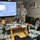 A U.S. military soldier in ACU (Army Combat Uniform) is conducting a briefing or training session for personnel wearing blue/grey digital camouflage uniforms, likely Iraqi or coalition security forces. The presentation is displayed on a projection screen and contains Arabic text with the title 'تطويق الموقع' (Cordon the Area/Site Containment). The setting appears to be a forward operating base or military compound briefing room.
