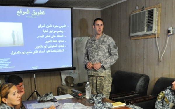 A U.S. military soldier in ACU (Army Combat Uniform) is conducting a briefing or training session for personnel wearing blue/grey digital camouflage uniforms, likely Iraqi or coalition security forces. The presentation is displayed on a projection screen and contains Arabic text with the title 'تطويق الموقع' (Cordon the Area/Site Containment). The setting appears to be a forward operating base or military compound briefing room.