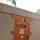 A rust-colored metal door is set into a concrete or stucco wall, marked with the acronym 'EOD' (Explosive Ordnance Disposal) and a black bomb silhouette above it. The door features a padlock hasp, a small window or label area, and decorative metal flanges on the sides suggesting a reinforced or themed enclosure. The setting appears to be an outdoor facility with gravel ground cover and trees visible in the background.