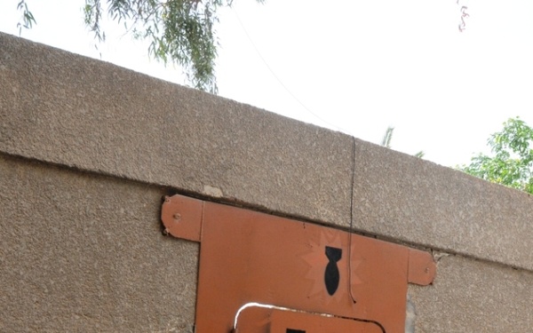 A rust-colored metal door is set into a concrete or stucco wall, marked with the acronym 'EOD' (Explosive Ordnance Disposal) and a black bomb silhouette above it. The door features a padlock hasp, a small window or label area, and decorative metal flanges on the sides suggesting a reinforced or themed enclosure. The setting appears to be an outdoor facility with gravel ground cover and trees visible in the background.
