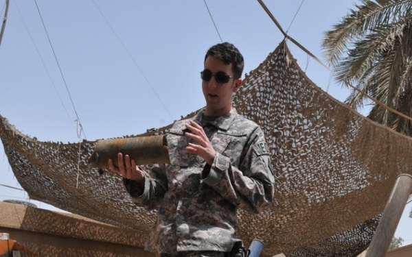 A U.S. Army soldier in ACU (Army Combat Uniform) stands in an outdoor area surrounded by various recovered ordnance and weapons components displayed on a gravel surface. The soldier appears to be conducting a briefing or demonstration, holding what appears to be a rocket or projectile component. The setting appears to be a forward operating base or military compound in a desert environment, likely used as an ordnance display or training area.