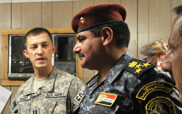 Two military/law enforcement personnel are shown in an indoor setting. A U.S. Army soldier identified by name tape as 'YANEY' stands alongside an Iraqi Federal Police commander wearing a dark blue digital camouflage uniform with a maroon beret. The Iraqi officer displays three-star rank insignia and a 'Federal Police Command' shoulder patch along with an Iraqi flag patch.