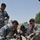 A U.S. Army EOD (Explosive Ordnance Disposal) soldier is conducting a training session with Iraqi security personnel wearing IED unit patches. The group is crouched on a gravel surface examining a small device or component on the ground. This appears to be a joint counter-IED training exercise in an outdoor military compound setting.