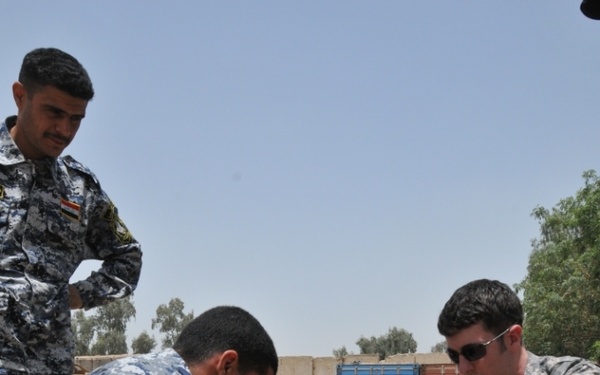 A U.S. Army EOD (Explosive Ordnance Disposal) soldier is conducting a training session with Iraqi security personnel wearing IED unit patches. The group is crouched on a gravel surface examining a small device or component on the ground. This appears to be a joint counter-IED training exercise in an outdoor military compound setting.
