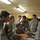 The image shows a group of personnel in what appears to be a training or briefing session inside a wooden-paneled room. Several individuals wearing blue-grey digital camouflage uniforms with Arabic script patches and Iraqi flag insignia are gathered around a grey equipment case on a table, while a person in US Army ACU (Army Combat Uniform) observes nearby. A civilian woman with sunglasses on her head appears to be acting as an interpreter or advisor.