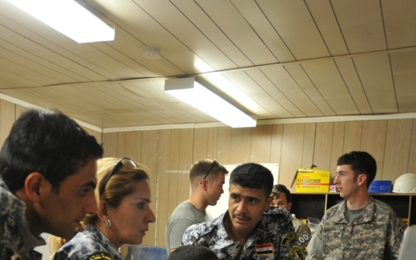 The image shows a group of personnel in what appears to be a training or briefing session inside a wooden-paneled room. Several individuals wearing blue-grey digital camouflage uniforms with Arabic script patches and Iraqi flag insignia are gathered around a grey equipment case on a table, while a person in US Army ACU (Army Combat Uniform) observes nearby. A civilian woman with sunglasses on her head appears to be acting as an interpreter or advisor.