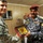 Two military personnel are engaged in a ceremonial exchange, with one individual in US Army uniform and another in Iraqi Federal Police uniform jointly holding a framed commemorative medallion or plaque. The setting appears to be a modest interior room with wood-paneled walls. This appears to be a formal gift-giving or recognition ceremony between coalition and Iraqi security forces.