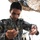 A military personnel wearing an EOD (Explosive Ordnance Disposal) uniform with Arabic script patches is examining wires connected to an assembly of plastic bottles and components under a camouflage net. The patch on the uniform reads 'EOD' in English and Arabic, indicating this is a bomb disposal or IED training/analysis context. The individual appears to be carefully inspecting or demonstrating an improvised device assembly.