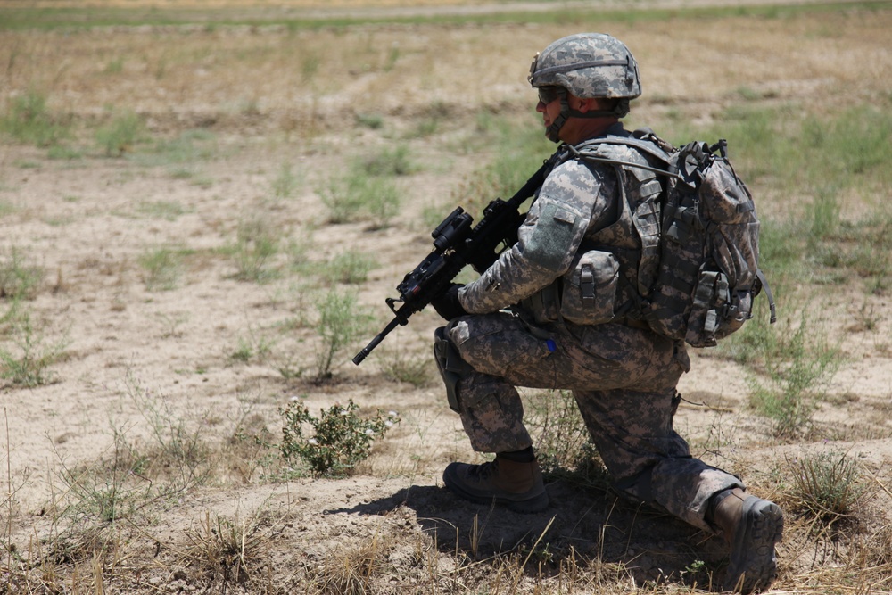 A soldier in full combat gear is kneeling on one knee in an arid, open field. The individual is wearing digital camouflage uniform (ACU pattern) and is holding an assault rifle while appearing to observe or scan the surrounding area. The setting suggests a field training exercise or operational patrol in a dry, semi-arid environment.