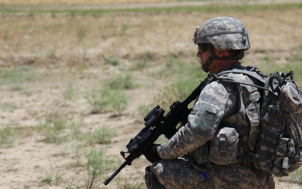 A soldier in full combat gear is kneeling on one knee in an arid, open field. The individual is wearing digital camouflage uniform (ACU pattern) and is holding an assault rifle while appearing to observe or scan the surrounding area. The setting suggests a field training exercise or operational patrol in a dry, semi-arid environment.