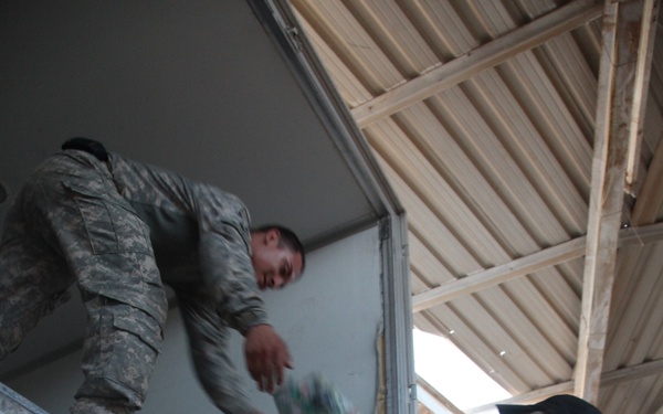 Two military personnel in U.S. Army uniforms are unloading or loading items from the back of a military truck or trailer under a covered structure. One soldier is positioned inside or on top of the truck bed, while the other stands on the ground passing or receiving an item. The setting appears to be a forward operating base or military logistics area.