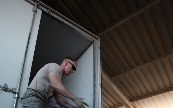 Military personnel in U.S. Army combat uniforms (ACUs) are unloading or loading a refrigerated truck/trailer inside a covered warehouse or storage facility. One soldier stands inside the truck bed passing items down to another soldier standing below. Additional personnel are visible observing or waiting nearby.