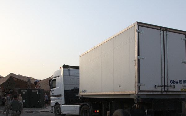 A white semi-truck with a box trailer marked 'Glow-R20-037' is entering or maneuvering through what appears to be a military base checkpoint or entry control point. Several soldiers in U.S. Army combat uniforms are present, monitoring the vehicle's movement. The setting includes concrete barriers, military tents, and a U.S. flag visible on a pole in the upper left.
