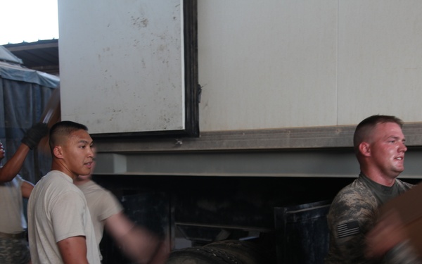 A group of military personnel in U.S. Army uniforms are engaged in a logistics or supply operation near a large white box truck or trailer. Soldiers are handling and moving boxes or cargo, with some in full ACU (Army Combat Uniform) and others in PT (physical training) attire. The scene appears to take place at a military installation or forward operating base.