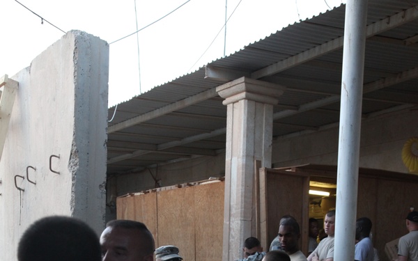 A group of military personnel in U.S. Army combat uniforms (ACUs) are engaged in a logistics or supply operation, passing and carrying cardboard boxes in what appears to be a partially covered outdoor area. The setting appears to be a forward operating base or similar military installation, possibly in a Middle Eastern environment based on the architecture and lighting. Multiple soldiers are visible in various stages of the box-passing chain.