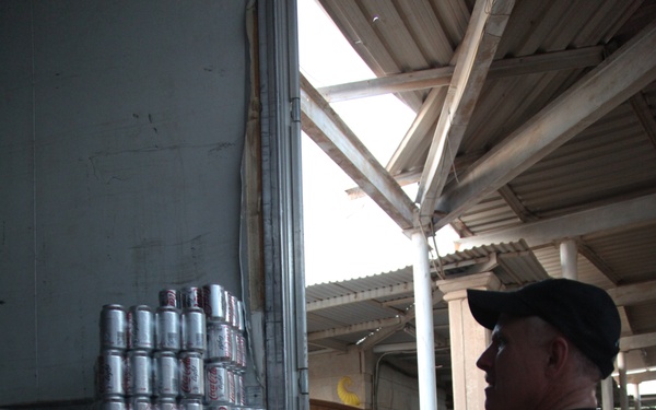 Military personnel in camouflage uniforms are unloading or distributing canned beverages, identifiable as Coca-Cola cans, from what appears to be a military vehicle or trailer. The scene takes place under a corrugated metal roof structure, suggesting a forward operating base or supply depot. Multiple soldiers are visible participating in the distribution activity.