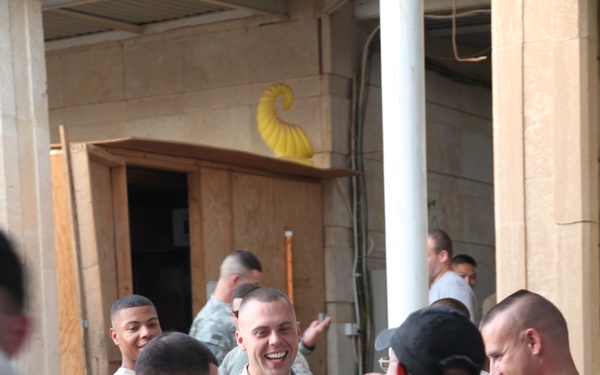 A group of U.S. Army soldiers in ACU (Army Combat Uniform) camouflage are engaged in what appears to be a work or supply detail at a forward operating base or military installation. The central soldier is smiling broadly while a package or box is being passed or tossed in the foreground. The setting appears to be an outdoor area adjacent to a concrete/stone building under some form of construction or renovation.