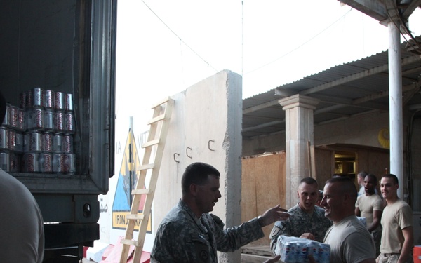 Military personnel in US Army combat uniforms are unloading or distributing supplies from a large truck in what appears to be a forward operating base or military installation. Soldiers are passing cases of bottled water and canned beverages, suggesting a resupply or distribution operation. The setting appears to be an arid, Middle Eastern environment based on the architecture and environmental conditions.