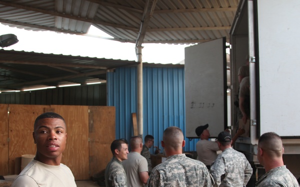 A group of military personnel in U.S. Army ACU (Army Combat Uniform) camouflage are engaged in a logistics or supply operation inside a covered warehouse or loading dock area. One soldier in the foreground faces the camera wearing a tan t-shirt and ACU trousers, while several others work near the open rear of a large white box truck. The setting appears to be a forward operating base or military installation.