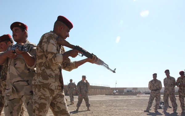 The image shows a military training exercise taking place in an arid, desert environment. Soldiers wearing desert camouflage uniforms and red berets are practicing weapons handling, with one soldier aiming an AK-pattern rifle while being assisted by another. Personnel in US Army uniforms are also present, suggesting a joint training or advisory scenario.