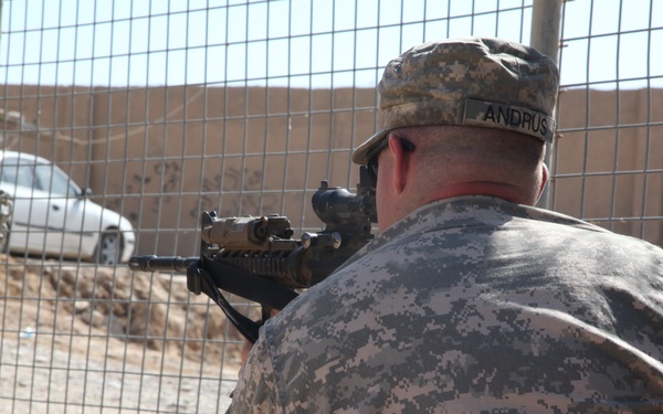 A U.S. Army soldier wearing Army Combat Uniform (ACU) digital camouflage is positioned behind a chain-link fence gate, aiming a rifle equipped with an optical scope. The name tape on the soldier's uniform reads 'ANDRUS'. The setting appears to be a forward operating base or checkpoint in an arid, desert environment.