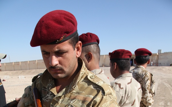 The image shows a group of military personnel standing in a line outdoors in an arid environment. The foreground soldier wears a distinctive tiger-stripe camouflage uniform and a dark red/maroon beret while holding an AK-pattern rifle. Other soldiers behind him wear desert camouflage uniforms and matching maroon berets.