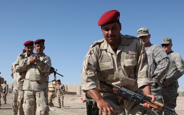 The image shows a military training exercise taking place in an arid, desert environment. Soldiers wearing tan/desert camouflage uniforms and red berets are practicing tactical movements while carrying AK-pattern rifles, observed by personnel in US Army ACU (Army Combat Uniform) pattern uniforms. The setting appears to be a training compound or base, suggesting an advise-and-assist or train-and-equip mission.