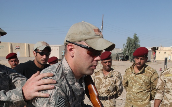 A U.S. Army soldier in ACU (Army Combat Uniform) with a '1st Infantry Division' patch and U.S. flag on his sleeve is demonstrating weapon handling with an AK-pattern rifle to a group of soldiers wearing red berets and desert camouflage uniforms. The setting appears to be an outdoor training area in an arid environment, consistent with a military advisory or training mission. A building in the background displays what appears to be an Iraqi flag.