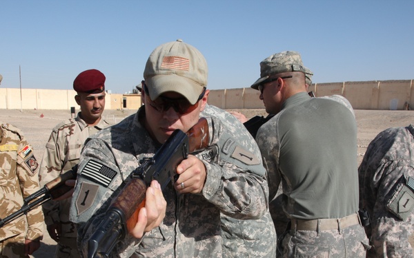The image shows a group of military personnel on what appears to be a training range in an arid environment. A soldier in US Army ACU (Army Combat Uniform) is handling an AK-pattern rifle, likely during a familiarization or training exercise. Personnel wearing different uniforms, including what appear to be Iraqi or Kurdish military uniforms, are observing.
