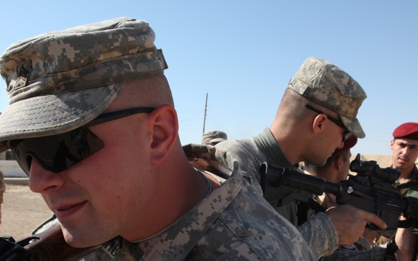 The image shows military personnel in a training or operational setting in what appears to be a Middle Eastern environment. Multiple soldiers wearing U.S. Army ACU (Army Combat Uniform) pattern camouflage are present alongside individuals in different uniforms, including one wearing a red beret. The soldiers are handling rifles in what appears to be a weapons handling or training exercise.