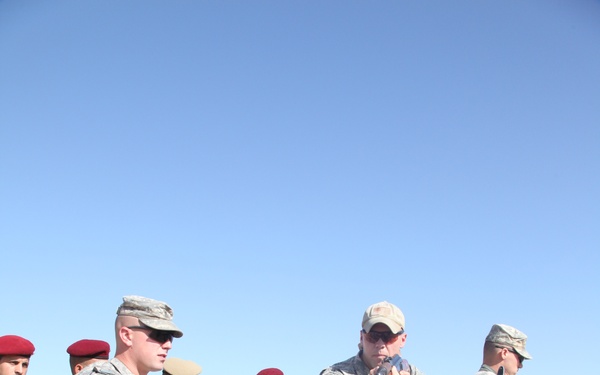 A group of military personnel in an arid outdoor environment appear to be engaged in a training exercise. Two soldiers in US Army ACU (Army Combat Uniform) pattern are demonstrating techniques, with one raising his leg in what appears to be a combatives or weapons-handling drill while another aims a pistol. Several other soldiers in tan/desert camouflage uniforms with maroon berets, consistent with Iraqi Army dress, observe the demonstration.