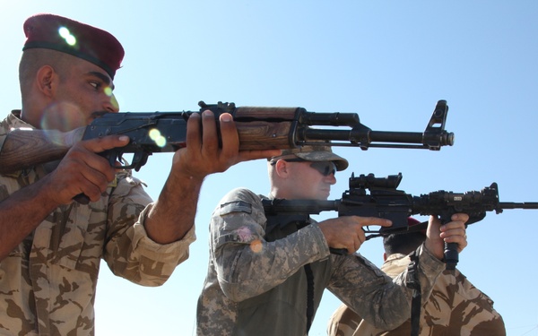 The image shows a military training exercise where soldiers in two different uniforms are practicing shooting stances side by side. Personnel in US Army ACU (Army Combat Uniform) pattern camouflage appear to be instructing or training alongside soldiers wearing a desert tan camouflage uniform with red berets. The setting appears to be an outdoor training range in an arid environment.