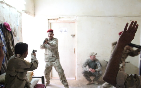 A group of military personnel in desert camouflage uniforms are gathered in a sparse interior room. Several individuals have their hands raised while one person appears to be filming or photographing with a small device. The atmosphere appears informal and social rather than tactical.