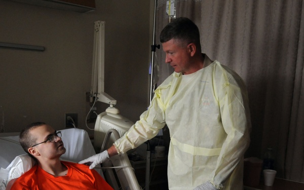 A hospital room scene showing a patient lying in a medical bed wearing a red shirt and dark pants, with visible IV lines and a hospital wristband. A standing individual wearing a yellow protective isolation gown and gloves is interacting with the patient, appearing to hold or check the patient's hand. The setting is a standard hospital room with medical equipment, curtains, and a bedside table visible.
